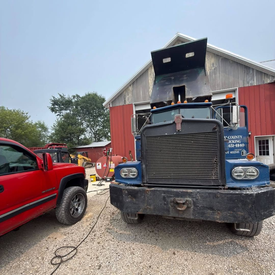A heavy dump truck parked at a farm shop next to the JT service pickup, with a welding lead run across the gravel